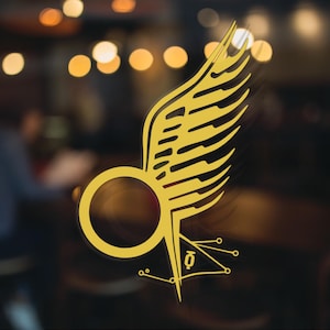 May include: A gold-colored winged symbol with a circular design in the center. The symbol is on a clear surface with a blurred background.