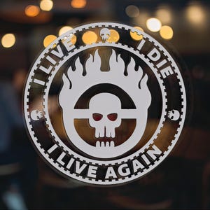 May include: A white and black circular sticker with a skull and flames in the center. The text around the skull reads "I live I die I live again".