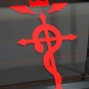 Może przedstawiać: Czerwona naklejka winylowa z "Symbolem Braci Flamel" z anime Fullmetal Alchemist. Symbol przedstawia koronę z skrzydłami, krzyż i węża.