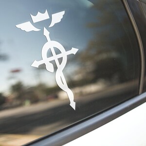 Może przedstawiać: Biała naklejka winylowa z symbolem Fullmetal Alchemist, przedstawiająca koronę z skrzydłami, krzyż i węża, naklejona na szybę samochodu.