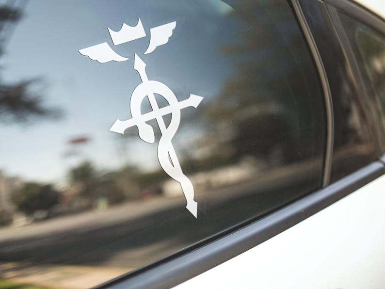 Może przedstawiać: Biała naklejka winylowa z symbolem Fullmetal Alchemist, przedstawiająca koronę z skrzydłami, krzyż i węża, naklejona na szybę samochodu.