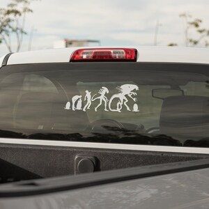 May include: A white truck with a rear window sticker depicting a humorous evolution of the Alien creature from the movie franchise. The sticker shows the creature evolving from a single-celled organism to its final form.