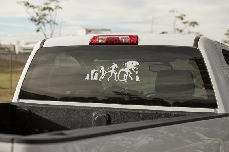 May include: A white truck with a rear window sticker depicting a humorous evolution of the Alien creature from the movie franchise. The sticker shows the creature evolving from a single-celled organism to its final form.