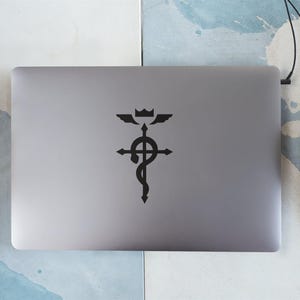 Może przedstawiać: Srebrny laptop z czarną naklejką z symbolem z anime Fullmetal Alchemist. Symbol to krzyż z wężem owiniętym wok&oacute;ł niego i koroną na szczycie.