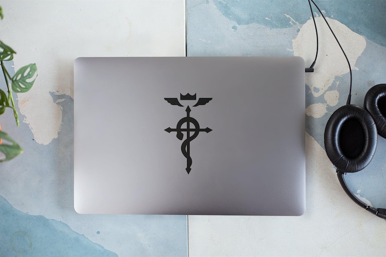 Może przedstawiać: Srebrny laptop z czarną naklejką z symbolem z anime Fullmetal Alchemist. Symbol to krzyż z wężem owiniętym wok&oacute;ł niego i koroną na szczycie.