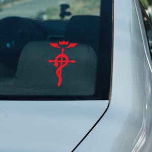 Może przedstawiać: Czerwony samoch&oacute;d z czerwoną naklejką z symbolem "Fullmetal Alchemist" na tylnej szybie.
