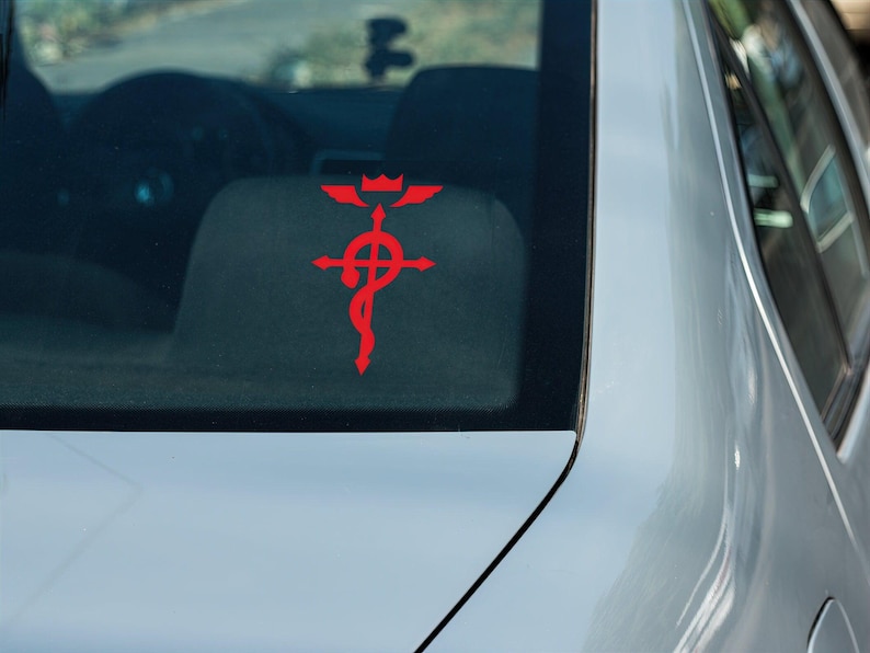 Może przedstawiać: Czerwony samoch&oacute;d z czerwoną naklejką z symbolem "Fullmetal Alchemist" na tylnej szybie.