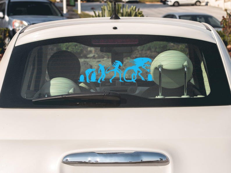 May include: A white car with a blue silhouette sticker on the back window. The sticker depicts a progression of alien-like figures, starting with a small, hunched figure and ending with a large, imposing figure.