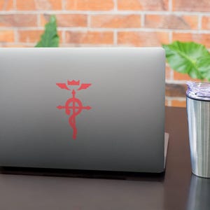 Może przedstawiać: Srebrny laptop z czerwonym symbolem na pokrywie. Symbol to krzyż z k&oacute;łkiem w środku i skrzydłami na g&oacute;rze. Laptop stoi na ciemnobrązowej powierzchni obok srebrnego kubka podr&oacute;żnego.