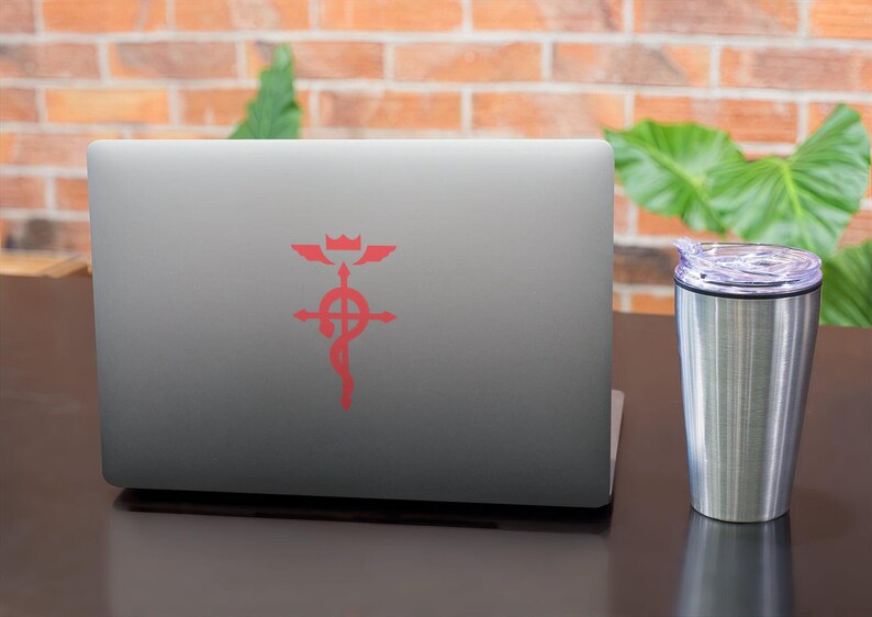 Może przedstawiać: Srebrny laptop z czerwonym symbolem na pokrywie. Symbol to krzyż z k&oacute;łkiem w środku i skrzydłami na g&oacute;rze. Laptop stoi na ciemnobrązowej powierzchni obok srebrnego kubka podr&oacute;żnego.