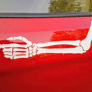 May include: A white skeleton hand pointing to the right, with the arm bent at the elbow, is on a red car.