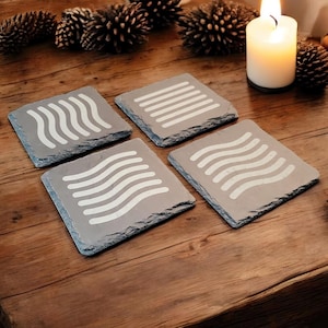 May include: Four gray slate coasters with white wavy lines designs. The coasters are arranged in a square pattern on a wooden surface.