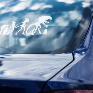 May include: A white vinyl sticker on the back window of a blue car. The sticker shows a humorous evolution of a creature from a simple organism to a Xenomorph from the Alien movie franchise.