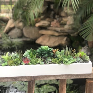 May include: A white rectangular planter filled with a variety of green succulents. The planter is sitting on a wooden railing.