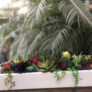 May include: A white rectangular planter box filled with a variety of succulents and greenery. The succulents are in shades of green, red, and purple. The planter is sitting on a wooden surface.