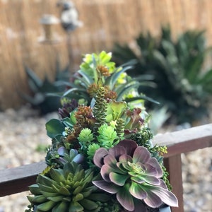 May include: A black planter filled with a variety of green and purple succulents. The succulents are arranged in a cascading pattern, with the largest succulents in the center of the planter.