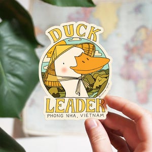 Op de afbeelding: Een sticker met een cartoon eend met een conische hoed. De eend heeft een oranje snavel en staat tegen een achtergrond van bergen en een blauwe lucht. De tekst luidt "DUCK LEADER" en "PHONG NHA, VIETNAM".