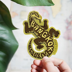 May include: A sticker featuring a stylized lizard with the word "BANGKOK" in brown text across its body. The sticker has a yellow-green border and the word "THAILAND" is written vertically along the tail. The background includes a map and green leaves.