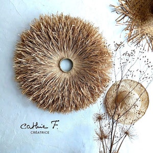 Può includere: Un grande arazzo da parete rotondo, intrecciato in modo naturale con paglia, con un'apertura circolare più piccola al centro. L'arazzo da parete è di colore beige e marrone e ha un aspetto soffice e testurizzato. L'arazzo da parete è esposto su una parete bianca con altri oggetti intrecciati naturali.