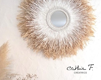 May include: Circular wall mirror with a natural, sunburst-style frame. The frame is made of layered, light-coloured, textured materials, surrounding a small, circular mirror. The mirror is mounted on a white wall. The text "Cathie F. Créatrice" is in the lower right corner.