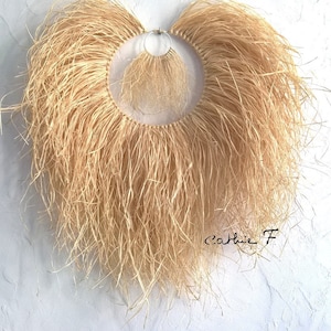 Puede incluir: Una gran pared colgante en forma de corazón hecha de paja natural con una pared colgante circular de paja más pequeña en el interior. La pared colgante más grande tiene una franja de largas y finas hebras de paja. La pared colgante más pequeña tiene una franja similar pero más corta. La pared colgante más grande está unida a la pared con un anillo de metal.