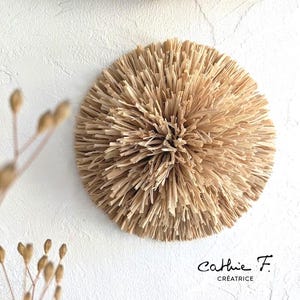 Pode incluir: Uma decoração de parede redonda feita de palha natural tecida, de cor castanho claro. A palha está disposta em forma radial, criando um aspeto texturizado e orgânico. A decoração é perfeita para adicionar um toque de beleza natural a qualquer divisão.