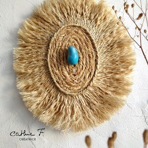 May include: A round wall hanging made of natural fibers with a fringe border. The center of the hanging features a woven circle with a turquoise blue stone in the middle. The text "Cathlie F. Creatrice" is visible at the bottom of the image.
