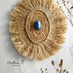 May include: A round wall hanging made of natural fibres with a fringe border. The centre of the hanging features a woven circle with a blue stone in the middle. The text "Cathie F. Creatrice" is visible at the bottom of the image.