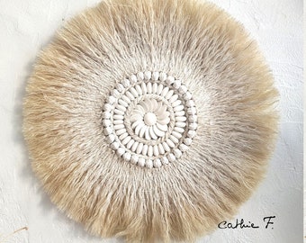 May include: A round wall hanging made of natural fibers with a white shell and bead design in the center. The design is surrounded by a fringe of natural fibers. The wall hanging is signed "Cathie F."