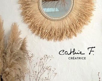 May include: A round wall mirror featuring a frame made of light tan natural fibers arranged in a sunburst pattern, with a beaded inner border. The text Cathie F. CRÉATRICE is printed in black on the right side of the mirror.