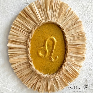 May include: A gold zodiac sign Leo wall decor with a straw-like frame. The sign is surrounded by a rope border and has a glittery gold background.