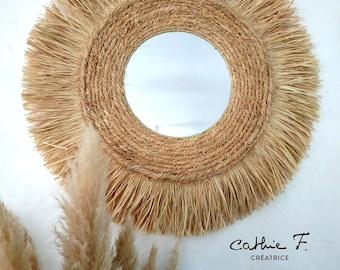 May include: A round mirror with a natural woven frame and a fringe of natural fibers. The mirror is hanging on a white wall with a bouquet of dried pampas grass in the foreground. The text "Cathie F. Creatrice" is visible in the bottom right corner of the image.