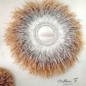 May include: A round mirror with a white and natural straw frame. The frame is made of woven straw and has a gradient of white to natural straw. The mirror is surrounded by a beaded border.