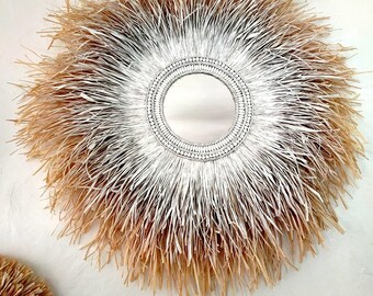 May include: A round mirror with a white and natural straw frame. The frame is made of woven straw and has a gradient of white to natural straw. The mirror is surrounded by a beaded border.