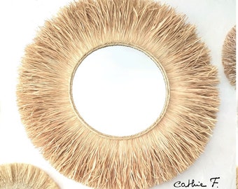 May include: A round mirror with a natural woven straw frame. The frame has a thick fringe of straw around the edge. The mirror is hanging on a white wall.