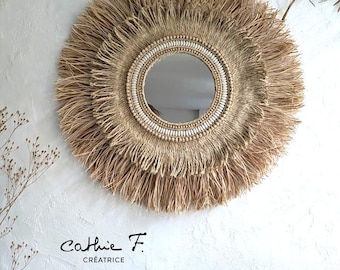 May include: A round mirror with a natural woven frame and white beads. The mirror is surrounded by a thick layer of natural fibers, creating a fringe effect. The text "Cathie F. Creatrice" is visible below the mirror.