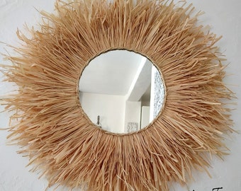 May include: A round mirror with a natural woven straw frame. The frame is thick and has a natural beige color. The mirror is reflecting a room with white walls.