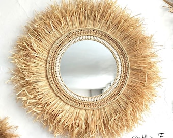 May include: Round wall mirror with a natural, sunburst design. The mirror features a central reflective surface surrounded by a frame of woven straw, wooden beads, and white shell accents. The outer edge has a fringe of straw. The mirror is a decorative home decor item.