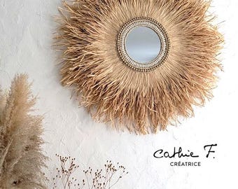 May include: Round wall mirror with a natural, sunburst-style frame made of dried grass. The mirror has a small, circular reflective surface and a decorative beaded border. The text "Cathie F. Créatrice" is in the lower right corner.