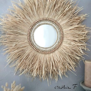 May include: A round mirror with a natural woven straw frame and a beaded border. The beads are in shades of beige and brown. The mirror is hanging on a grey wall.