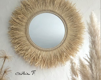 May include: A round mirror with a natural woven frame. The frame is made of straw-like material and has a fringe around the edge. The mirror is hanging on a white wall.
