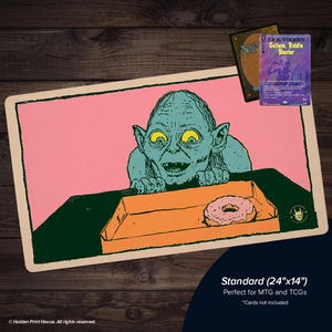 Puede incluir: Un tapete de juego rectangular con una criatura de piel verde azulado y grandes ojos amarillos mirando un dónut glaseado rosa en una caja naranja. El fondo es rosa con un borde verde oscuro. Una carta coleccionable de Gollum se sitúa encima.