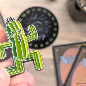 May include: A green and yellow enamel pin featuring a cartoon cactus character with arms raised. The pin has a gold outline and is set against a blurred background with a black circular object and trading cards.