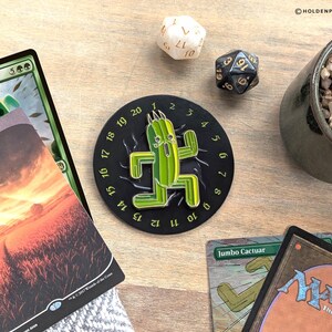 May include: A black and green Cactuar-themed game accessory with a time-telling design, featuring a green cactus figure. The image includes Magic: The Gathering cards, dice, and a small potted plant, all arranged on a wooden surface.