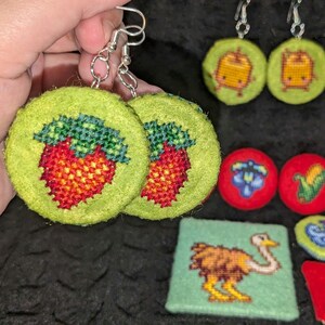 May include: A pair of green felt earrings with a red and green embroidered strawberry design. The earrings are hanging from silver hooks.