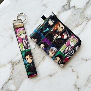 May include: A fabric keychain and a matching rectangular zippered pouch featuring a multi-panel print of various anime characters with distinct hair colors and facial expressions. The keychain has a silver metal ring and a flat rectangular connector.