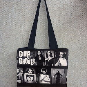 Puede incluir: Bolso tote negro con un collage de portadas de c&oacute;mics de terror vintage e im&aacute;genes de monstruos de pel&iacute;culas cl&aacute;sicas. El bolso tiene un asa negra y las palabras "Girls Will Be Ghouls" impresas.