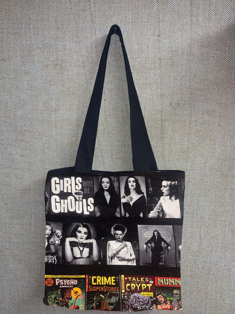 Puede incluir: Bolso tote negro con un collage de portadas de c&oacute;mics de terror vintage e im&aacute;genes de monstruos de pel&iacute;culas cl&aacute;sicas. El bolso tiene un asa negra y las palabras "Girls Will Be Ghouls" impresas.