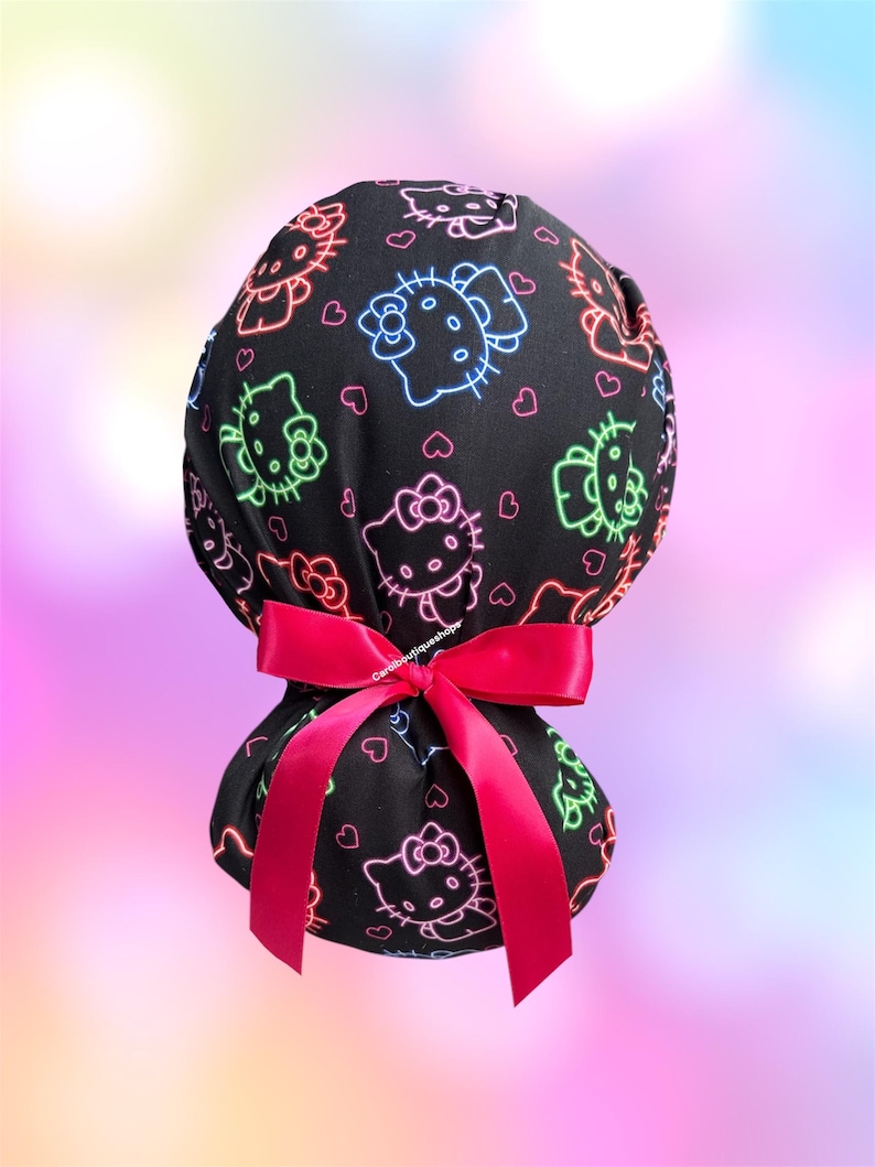 May include: A black scrub cap featuring a neon-style print of Hello Kitty characters in blue, green, and pink outlines, interspersed with small pink hearts. The cap is gathered at the back and secured with a bright pink satin ribbon bow.