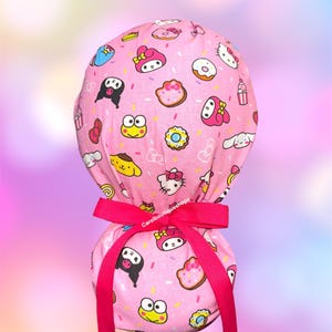 May include: A pink scrub cap featuring a repeating pattern of Sanrio characters including Hello Kitty, My Melody, Kuromi, Keroppi, and Pompompurin, interspersed with donuts and sprinkles. The cap is secured with a bright pink grosgrain ribbon.